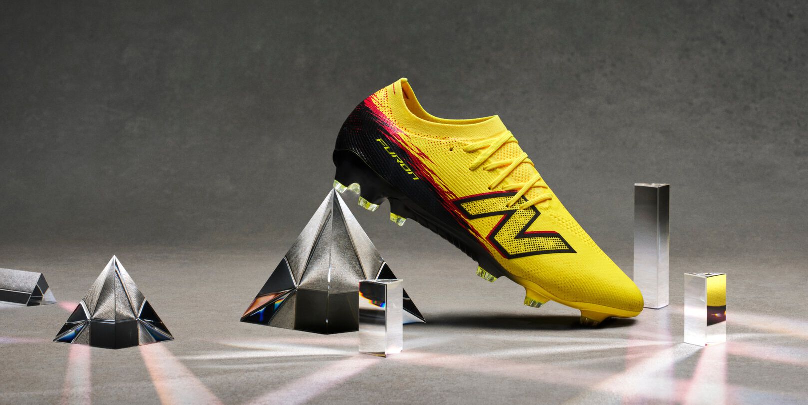A bright yellow soccer cleat with black and red accents is displayed on its side among several glass prisms on a neutral gray surface. 